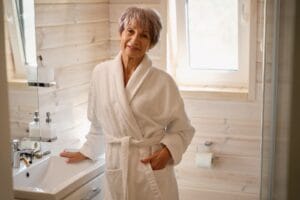 An older adult woman is standing in a bathroom, wearing a bathrobe. She smiles at the camera.