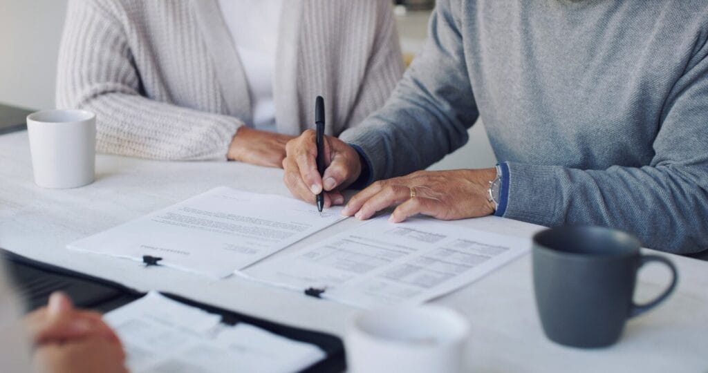 Close-up of a desk with paperwork and three coffee mugs. An older adult man's hands are signing the paperwork. There is a woman seated next to him and a person across the desk from him.