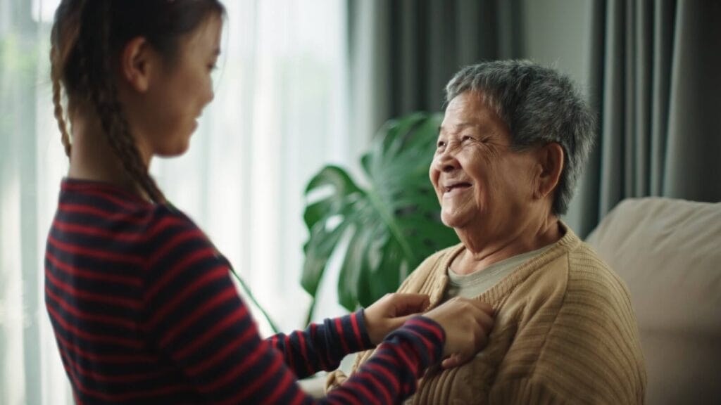 A woman helps an older adult woman put on a cardigan. They are smiling.