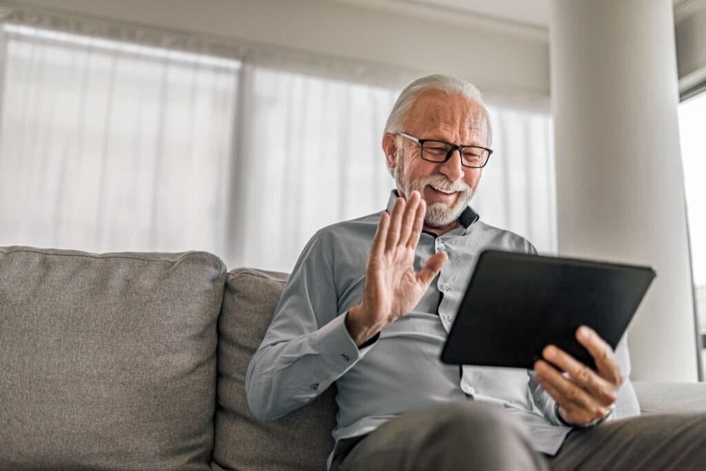 An older adult man is sitting on his couch, holding a tablet device, and waving at someone he is video chatting with.