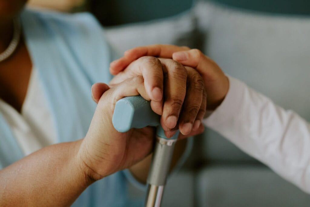 Close-up of an older adult woman's hands resting on the handle of a cane. Another person's hand is on top of hers.