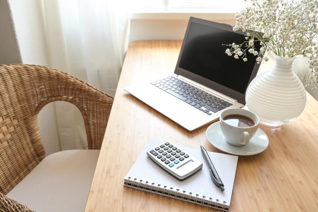 Image of a person's home office desk, with a laptop, a calculator, a notebook, a pen, a cup of coffee, and a vase with baby's breath flowers in it.
