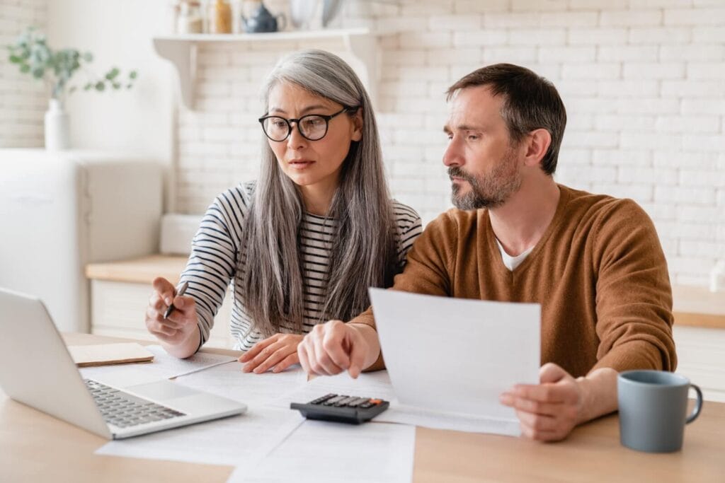 A woman and a man sit at a kitchen table. They are looking over paperwork. There is a laptop in front of the woman and she is pointing to it.