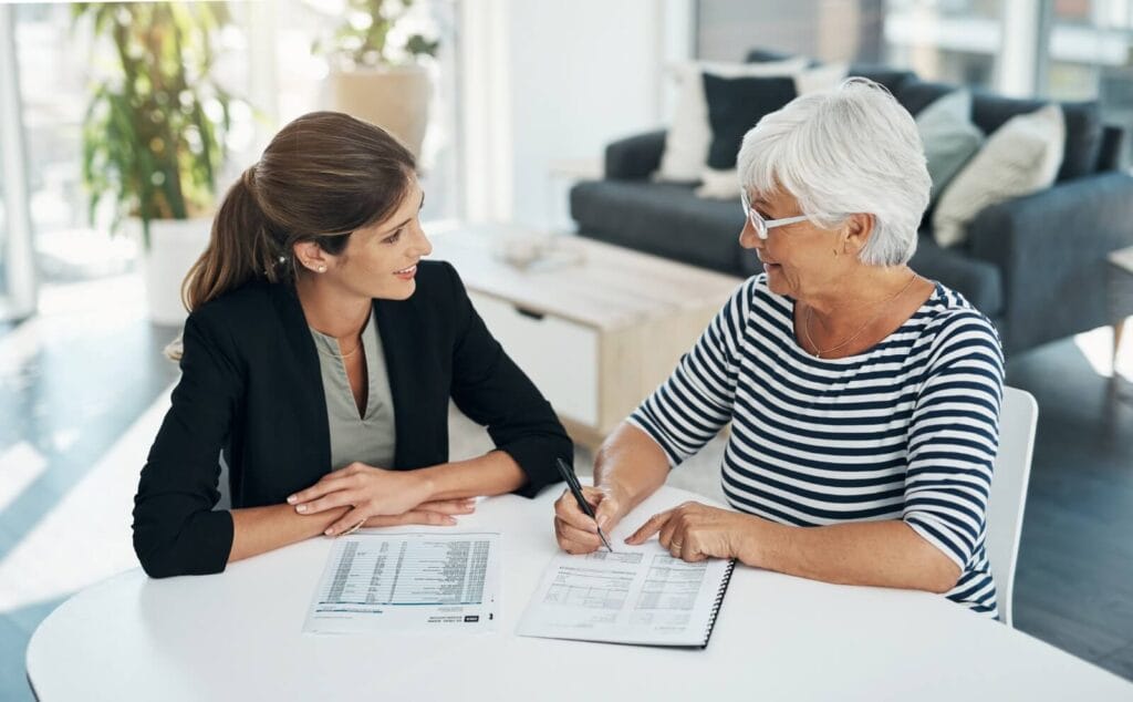 A woman and an older adult woman are sitting at a table with paperwork in front of them. The older woman is holding a pen to sign one of the documents.