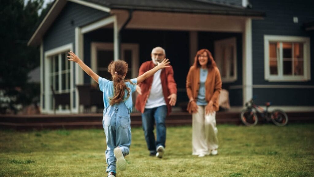 An older adult man and woman are walking in front of their house to greet their granddaughter, a young girl who is running toward them with her arms in the air.