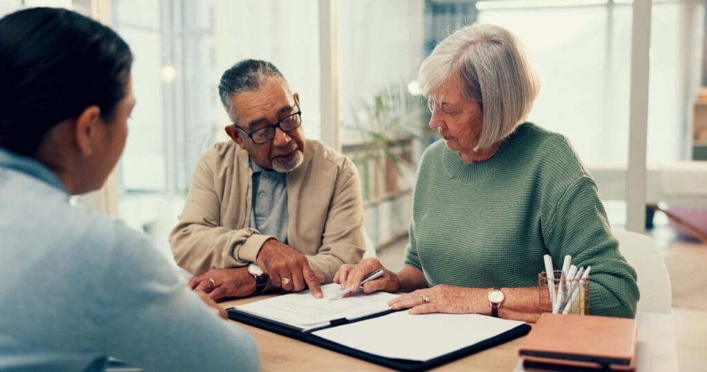 An older adult man and woman are sitting at a desk across from a man. They are looking over paperwork, and the woman is holding a pen.