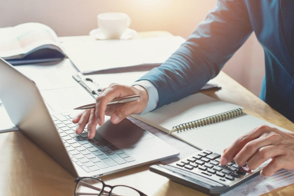 Image of a desktop, with a person's hands typing on a laptop and using a calculator. There is also a notebook, some paperwork, a pair of glasses, and a cup of coffee on the desk.