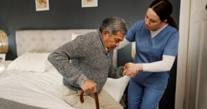 A woman care aide helps an older adult man stand up from a bed. He is using a cane.