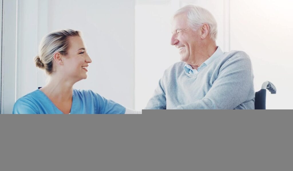 A female caregiver crouches beside an older adult man who is seated in a wheelchair. They are holding hands and smiling at each other.
