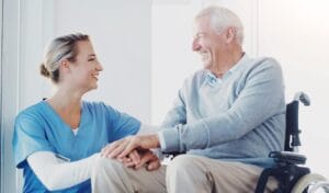 A female caregiver crouches beside an older adult man who is seated in a wheelchair. They are holding hands and smiling at each other.
