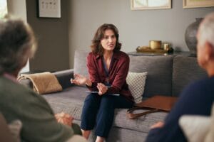A woman holding some papers and sitting on a couch is talking to an older adult man and woman who are seated in the foreground with their backs to the camera.