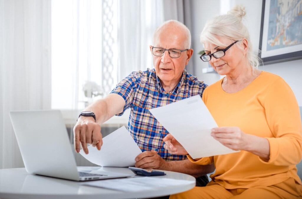 An older adult man and woman are seated at a table with a laptop and a calculator on it. They are holding papers, and the man is pointing to the laptop.