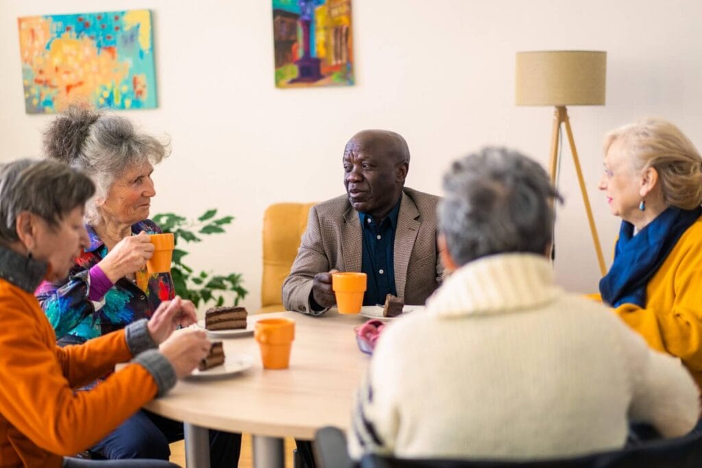 A group of four older adult women and one older adult man are sitting at a table together, eating cake and drinking coffee.