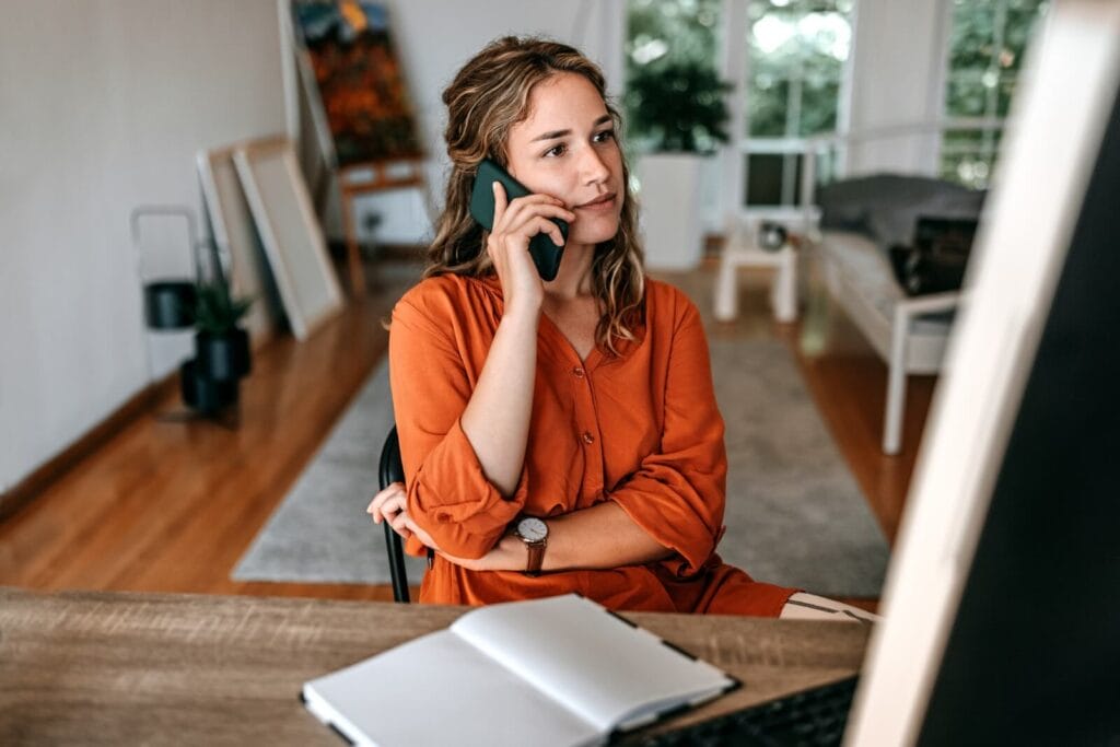 A woman is speaking on her cell phone. She is seated at a desk with a notebook in front of her.
