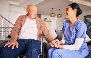 An older adult man is sitting in a wheelchair, holding the hands of a woman caregiver who is sitting on the couch next to him.