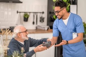A male professional caregiver hands a bowl to an older adult man, who is seated at his kitchen table.