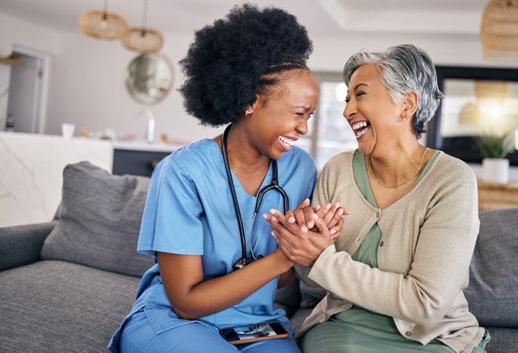 A female home care aide sits on a couch with an older adult woman. They are smiling at each other and holding each other's hands.