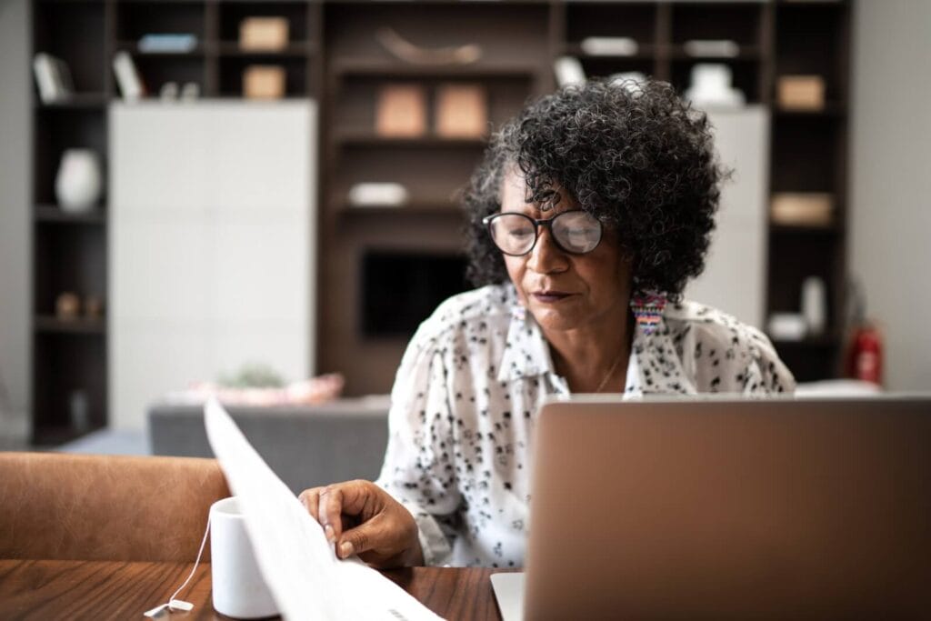 An older adult woman sits at a table with a laptop, paperwork, and a cup of tea in front of her.
