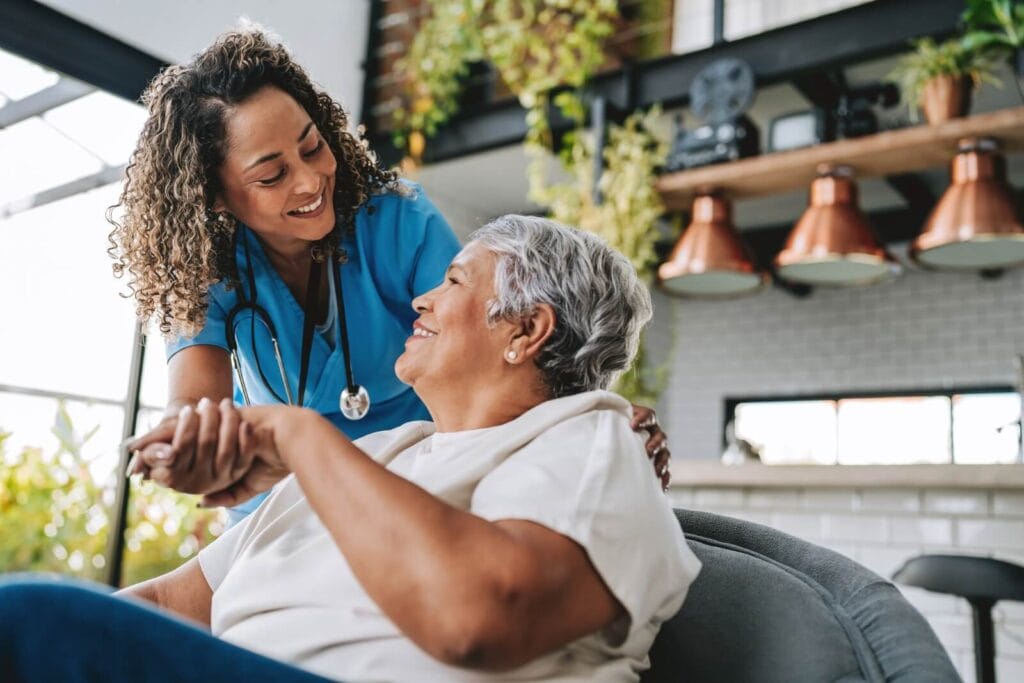 An older adult woman sits in a chair, looking up at a woman health care worker, who is holding her hand.