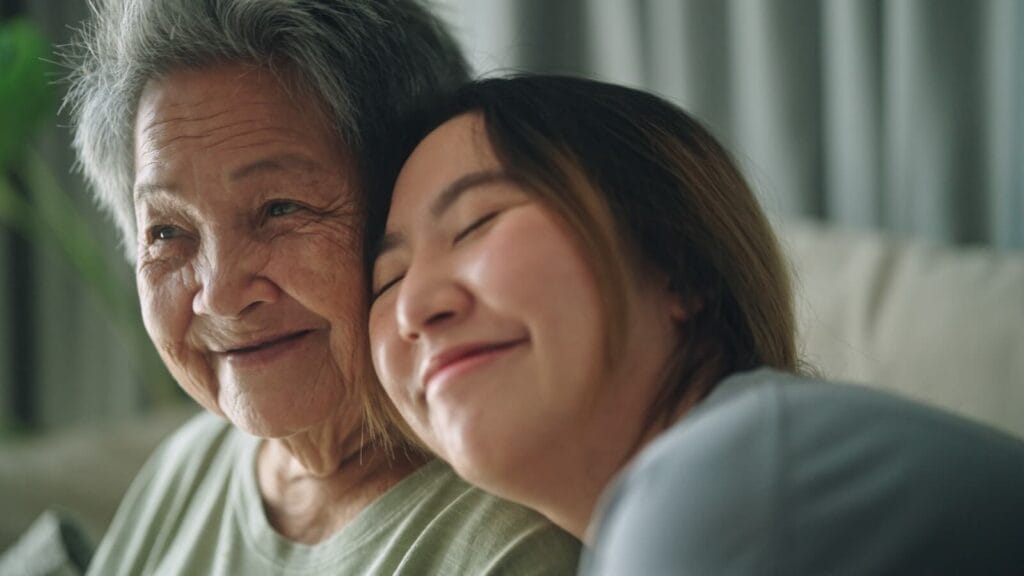 A woman is resting her head on her older adult mother's shoulder. They are smiling.