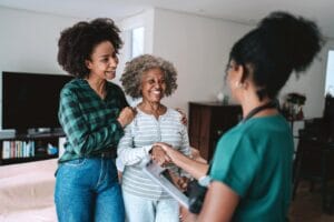 A woman and her older adult mother stand in a living room, talking to a woman home care aide. The older adult woman shakes the aide's hand, and everyone is smiling.