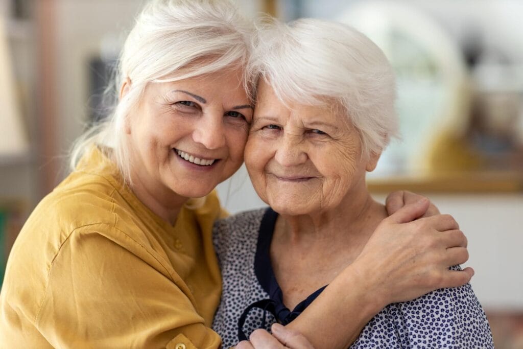 A woman and her older adult mother are hugging and smiling at the camera.