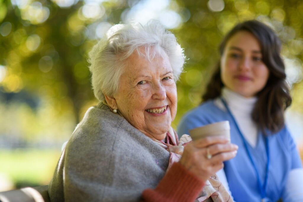An older adult woman sits outside, holding a cup and smiling at the camera. There is a woman professional caregiver sitting next to her in the background.