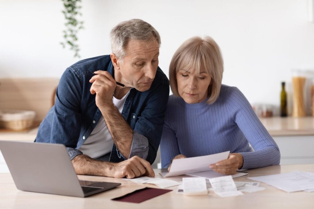 An older adult man and woman site at a table with papers, receipts, and a laptop in front of them.