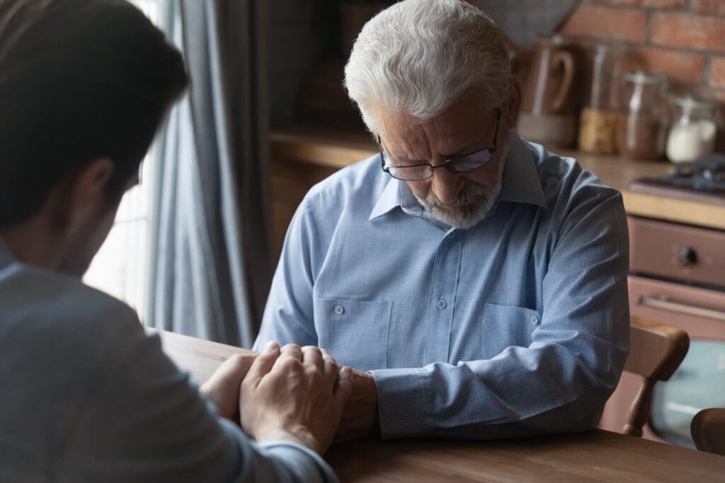 A man sits across a table from his older adult father, holding his hands. The father looks down, appearing sad.
