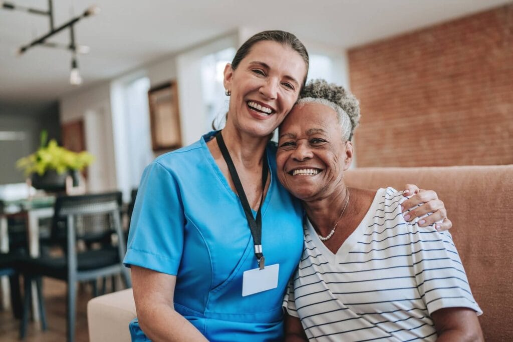 A woman home care aide sits with an older adult woman on a couch. They have their arms around each other and are smiling.