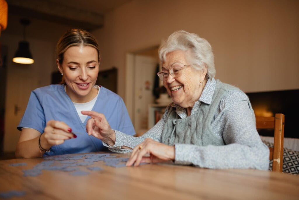 A woman caregiver and an older adult woman sit at a table, working on a jigsaw puzzle.