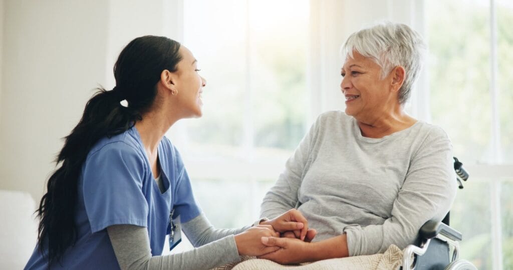 A woman caregiver crouches next to an older adult woman seated in a wheelchair, holding her hands.