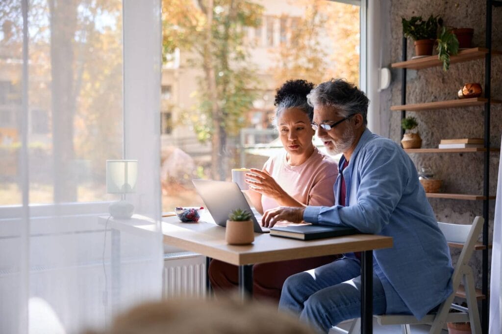 An older adult man and woman sit at a desk looking at a laptop in front of them,.