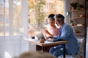 An older adult man and woman sit at a desk looking at a laptop in front of them,.