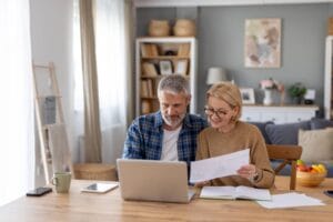 An older adult man and woman sit at a table with a laptop, a notebook, and some paperwork in front of them.
