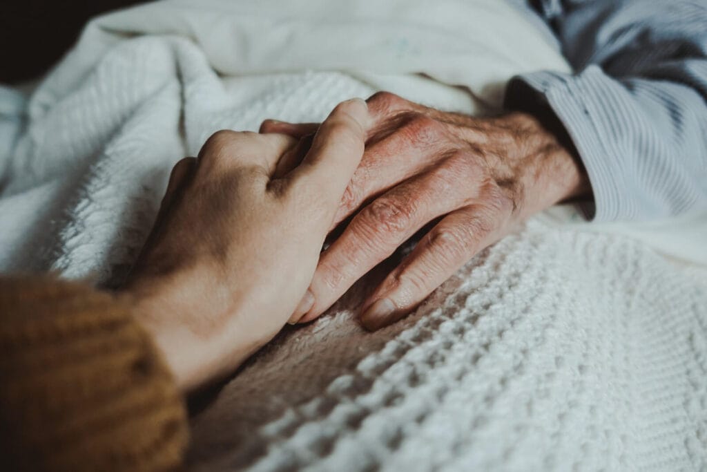 A close-up of a woman's hand holder an older adult's hand over a blanket.