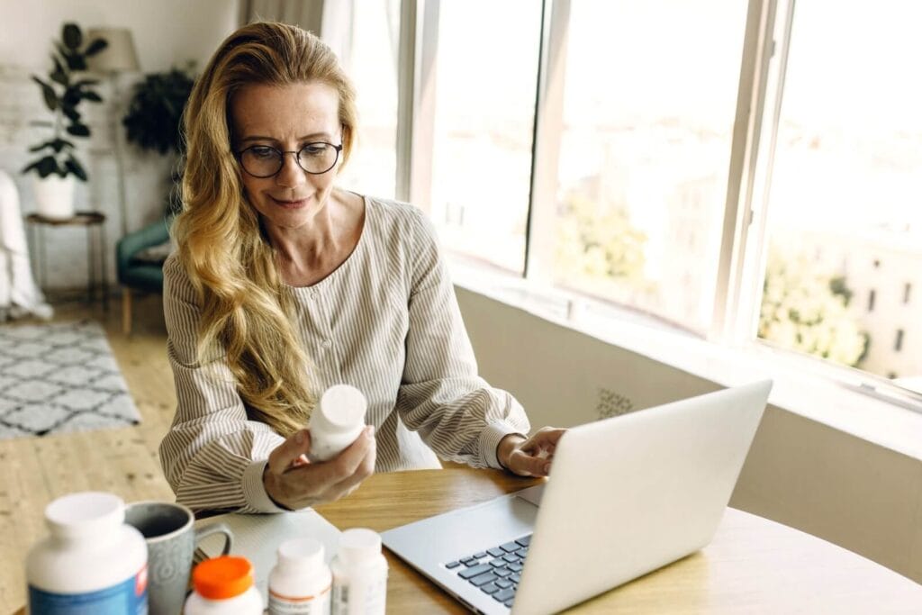 An older woman is holding a bottle of medication and working on a laptop computer.