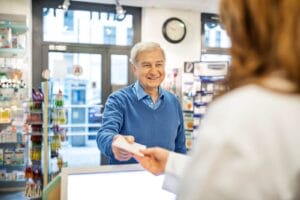 An older adult man hands a prescription to a pharmacist.