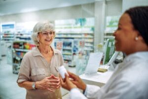 An older adult woman is standing at a pharmacist's window. The woman pharmacist is holding a bottle of medication.