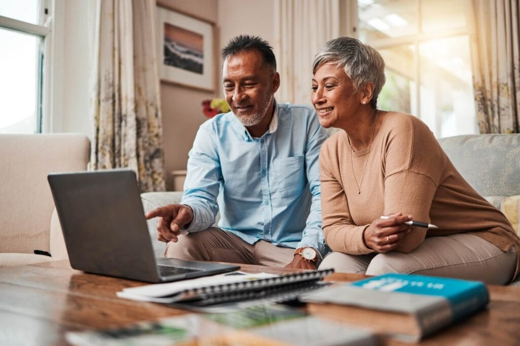 A mature woman and man sit on a couch, looking at a laptop on a table in front of them. There are notebooks on the table, too.