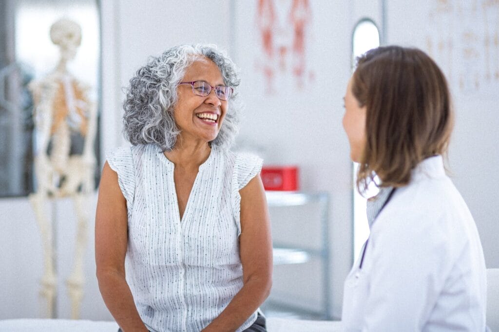 An older adult woman and a woman doctor sit in a doctor's office, talking and smiling.