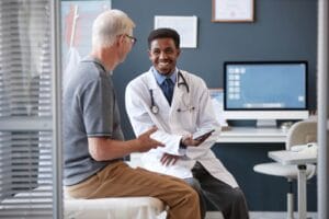 An older adult man sits on a examination table next to a younger male doctor. They are smiling.