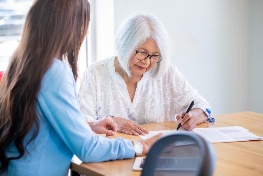 An older adult woman is sitting at a table with a younger woman, signing some paperwork.