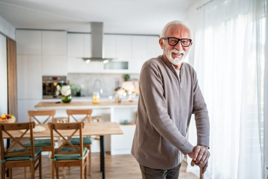 An older adult man holding a cane stands in a living area of his home. Behind him is the kitchen area with a dining table.