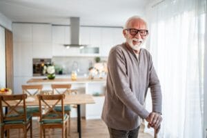 An older adult man holding a cane stands in a living area of his home. Behind him is the kitchen area with a dining table.