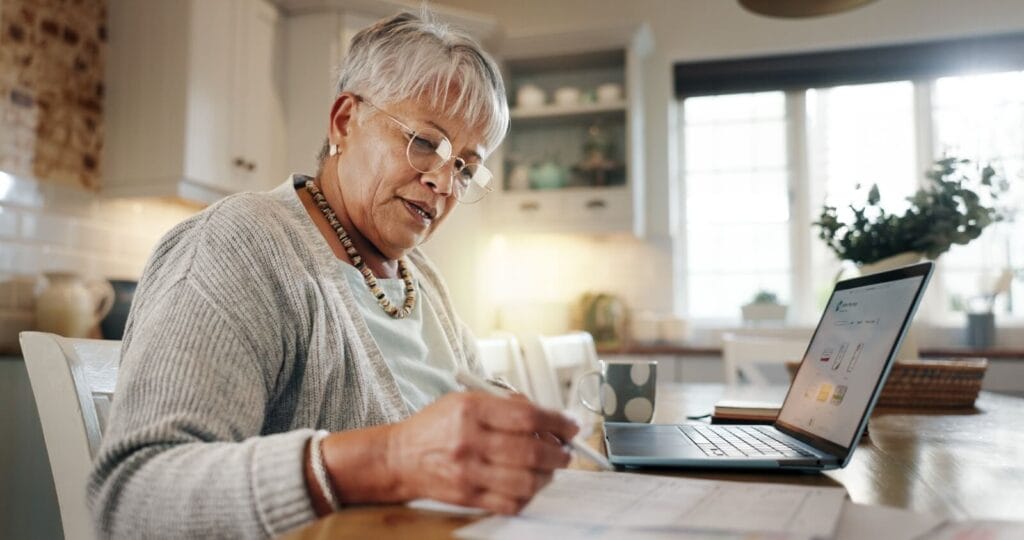 An older adult woman sits at a table with a laptop and paperwork in front of her.