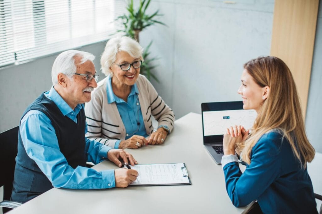 An older adult man and woman sit at a desk with a woman. The man has a clipboard in front of him and the younger woman has a laptop.