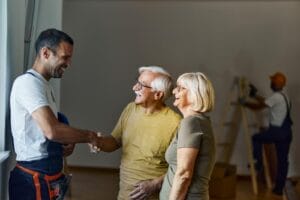 A contractor shakes the hand of an older adult man who is standing next to his wife. There is a man climbing a ladder and wearing a hardhat in the background.