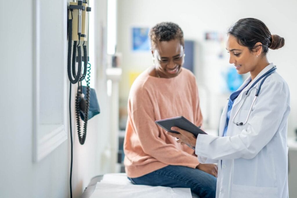 An older adult woman sits on a doctor's examination table, speaking to a younger woman doctor. The doctor is holding a tablet, showing the older woman something.