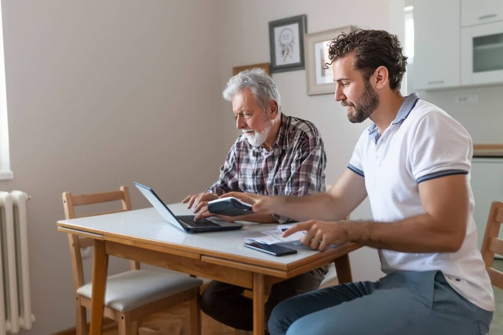 A man and his older adult father sit at a table with paperwork, a calculator, and a laptop in front of them.
